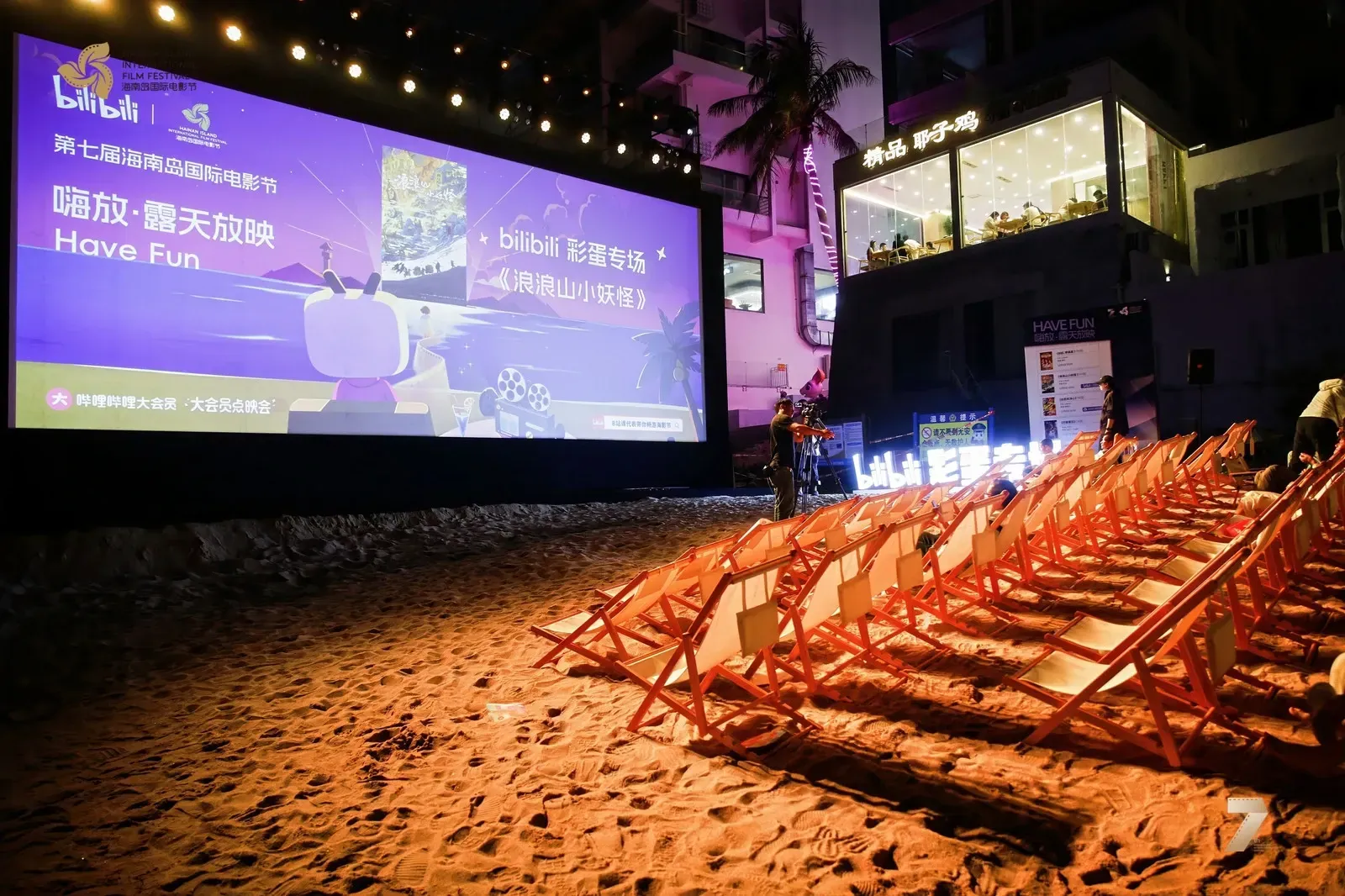 An open-air beach screening in Tianya Town at the 7th HIIFF, with beach loungers in front of the screen and coconut palms and street buildings behind